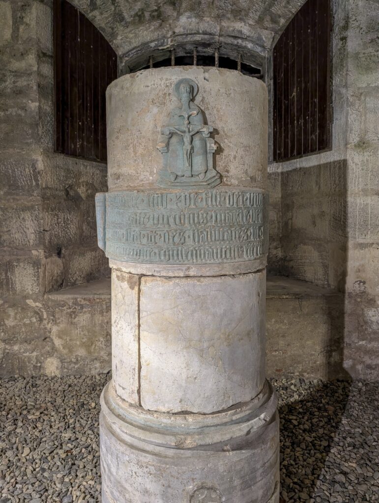 Steinsäule im gewölbten Durchgang der Bastion, oben ein grünlich gefasstes Relief der Dreifaltigkeit, darunter eine Schriftkartusche in gotischen Minuskeln