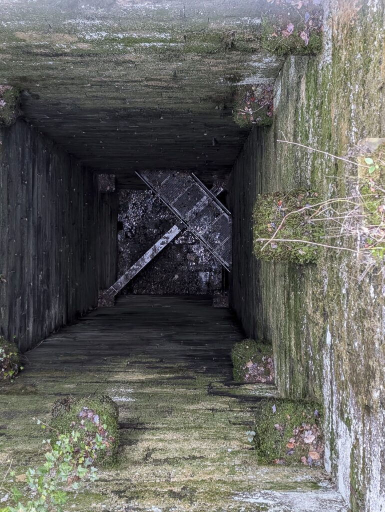 Blick senkrecht in den tiefen, gemauerten Brunnenschacht mit einer diagonalen Metallbrücke